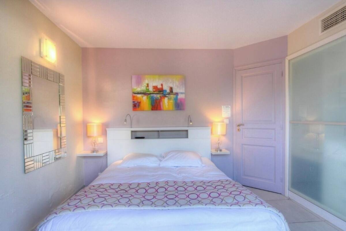 Une chambre à coucher avec un lit, une table de chevet, un miroir fixé au mur, un tableau et une porte.