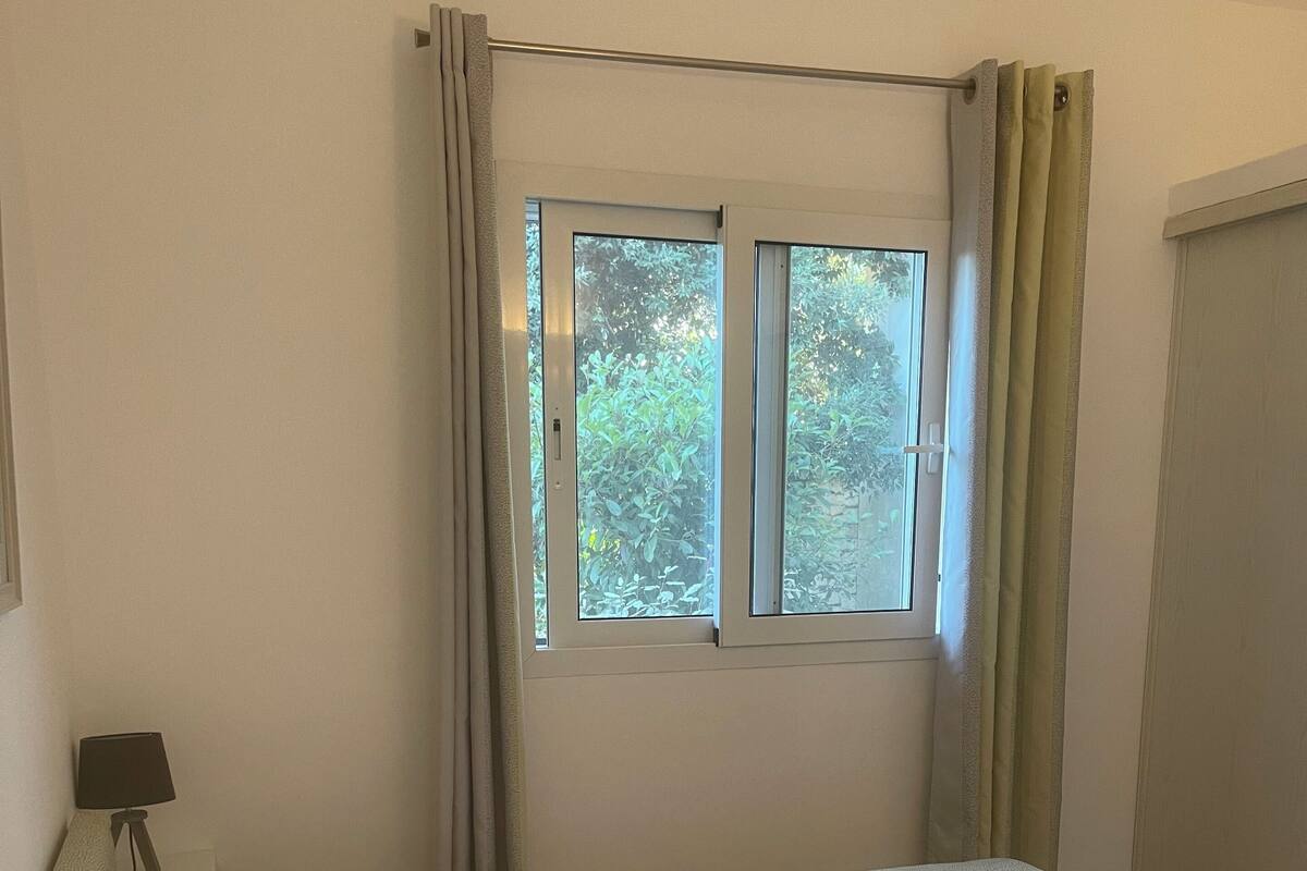 Une chambre d’hôtel avec un lit, une table de chevet avec une lampe, une fenêtre avec des rideaux et une vue sur la verdure.