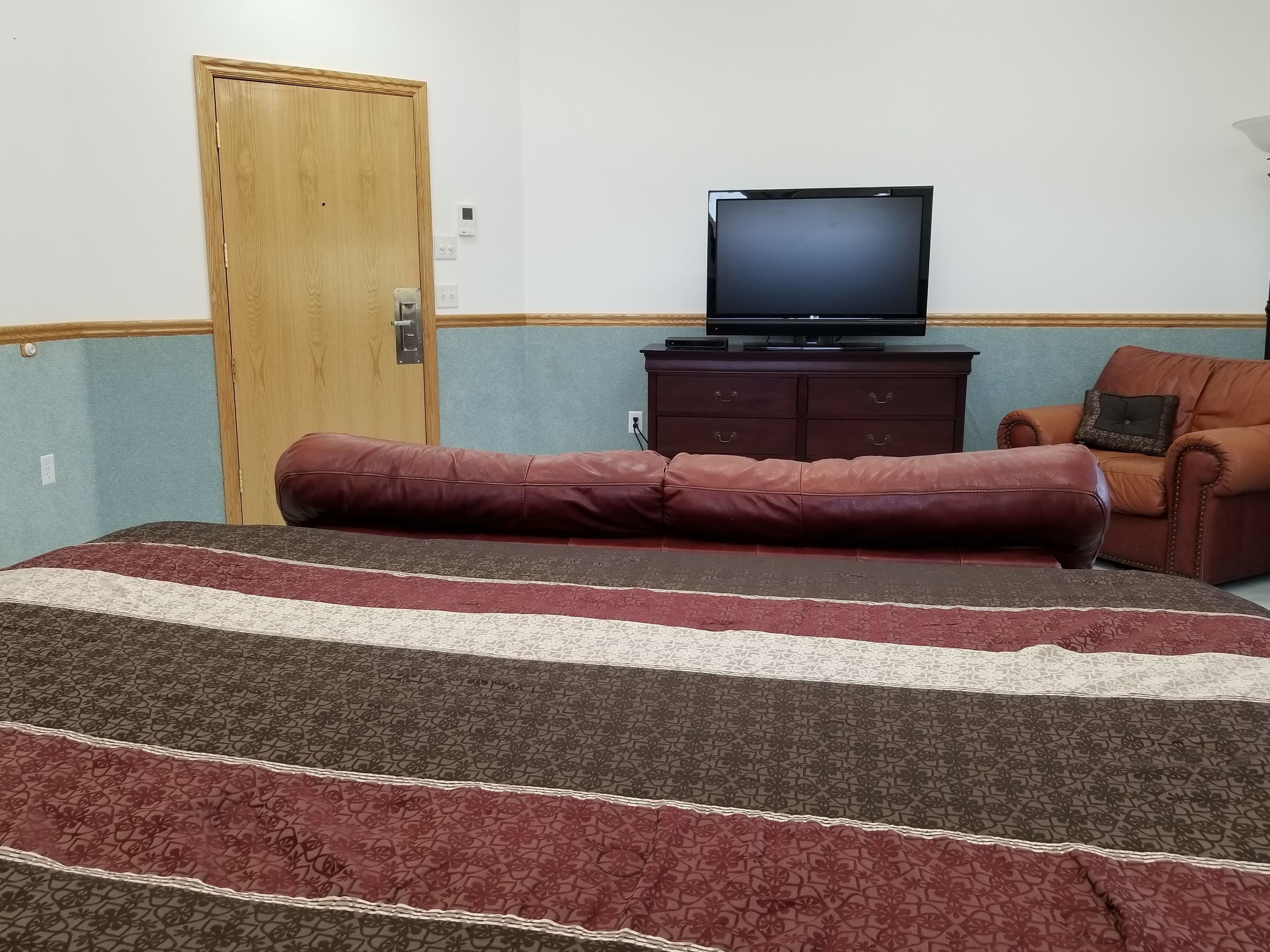 A room with a bed, a television on a stand, a wooden door, and a brown leather couch.