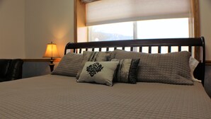 A bed with a quilted bedspread and multiple pillows, a nightstand with a lamp, and a window with a view of mountains. - Zion's Most Wanted Hotel (Hildale)