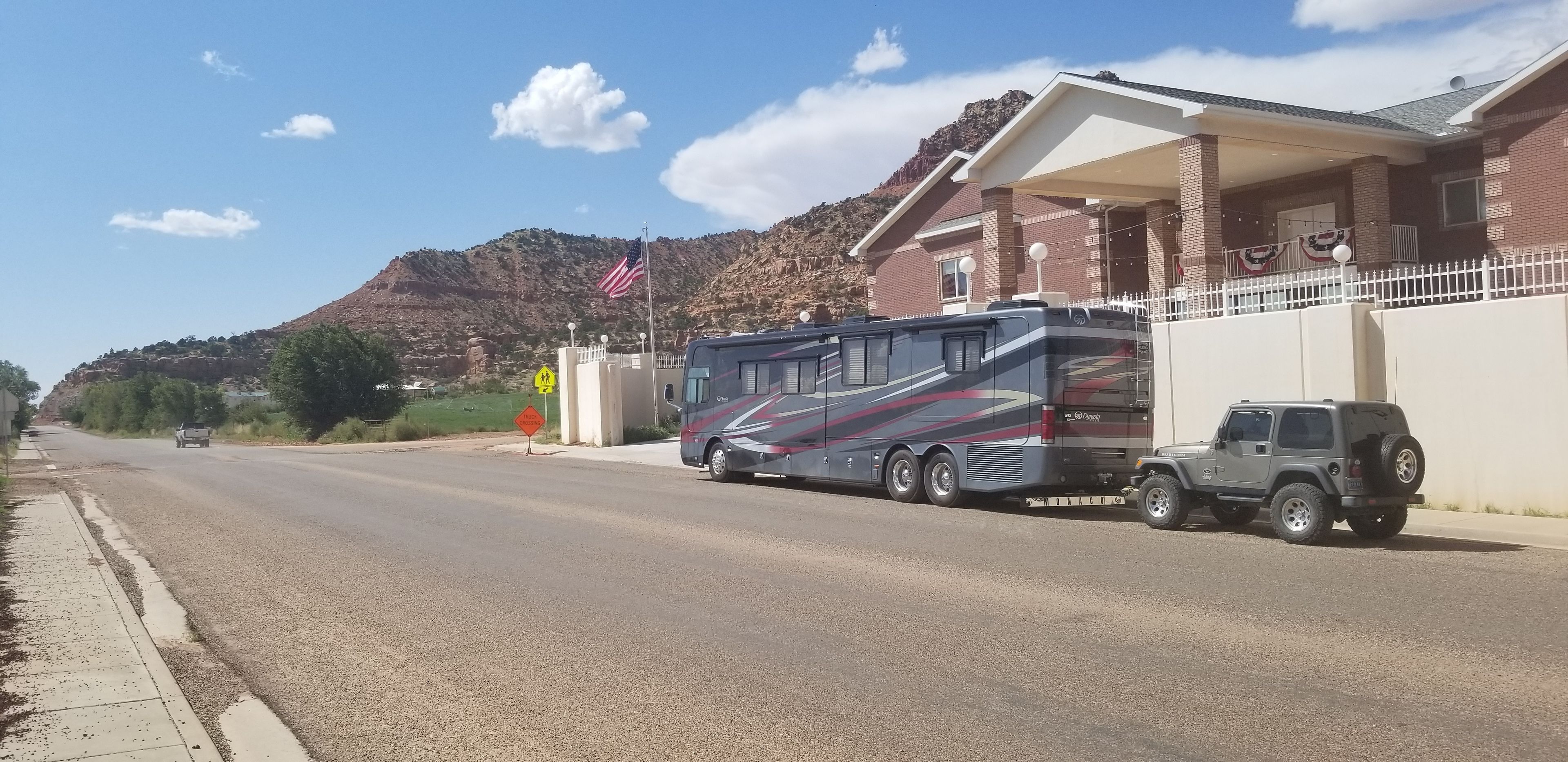 A large RV parked in front of a building with a flag, a Jeep, and a road with a mountain in the background.