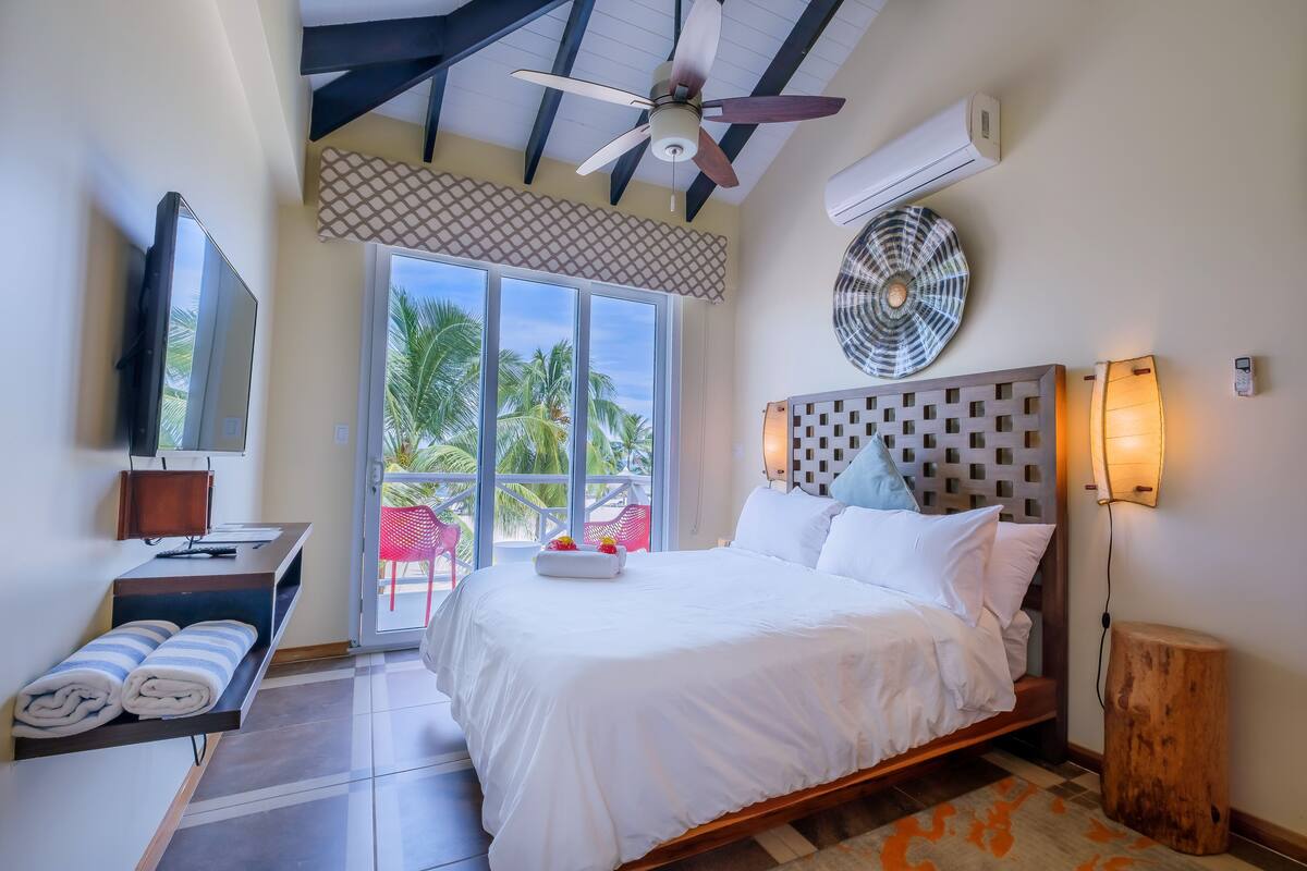 A hotel room with a bed, a desk with a TV, a ceiling fan, and a view of palm trees.