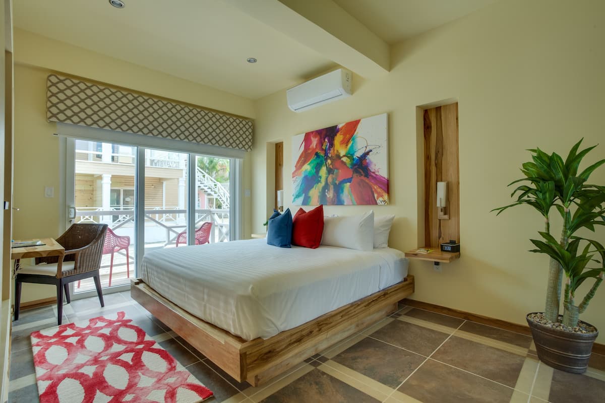A bedroom with a bed, a desk, a chair, a potted plant, and a colorful abstract painting on the wall.