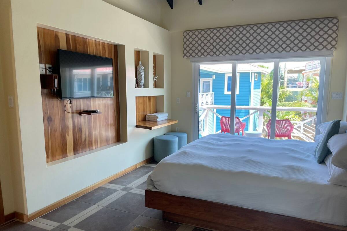 A bedroom with a large bed, a TV mounted on a wooden panel, a small seating area, and a balcony with a view of buildings and greenery.