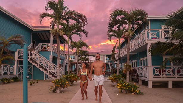 A couple walking on a sandy path between two buildings with balconies, surrounded by palm trees and flowers. - Brisa Oceano Resort (Placencia)