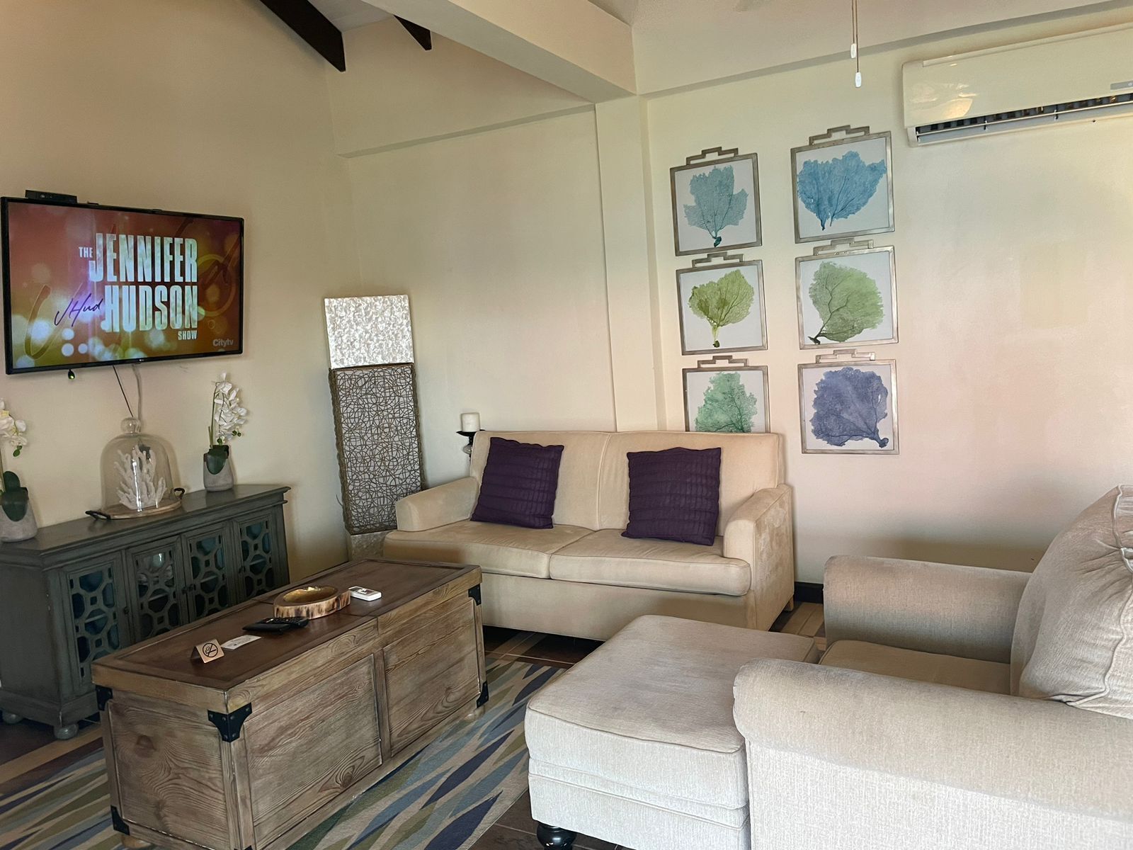 A living room with a beige sofa, a wooden coffee table, and a TV displaying 'The Jennifer Hudson Show'. There are framed botanical prints on the wall and a decorative piece above the sofa.