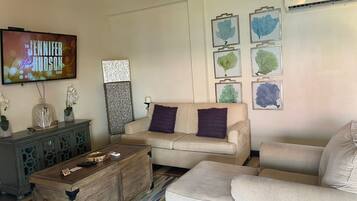 A living room with a beige sofa, a wooden coffee table, and a TV displaying 'The Jennifer Hudson Show'. There are framed botanical prints on the wall and a decorative piece above the sofa.