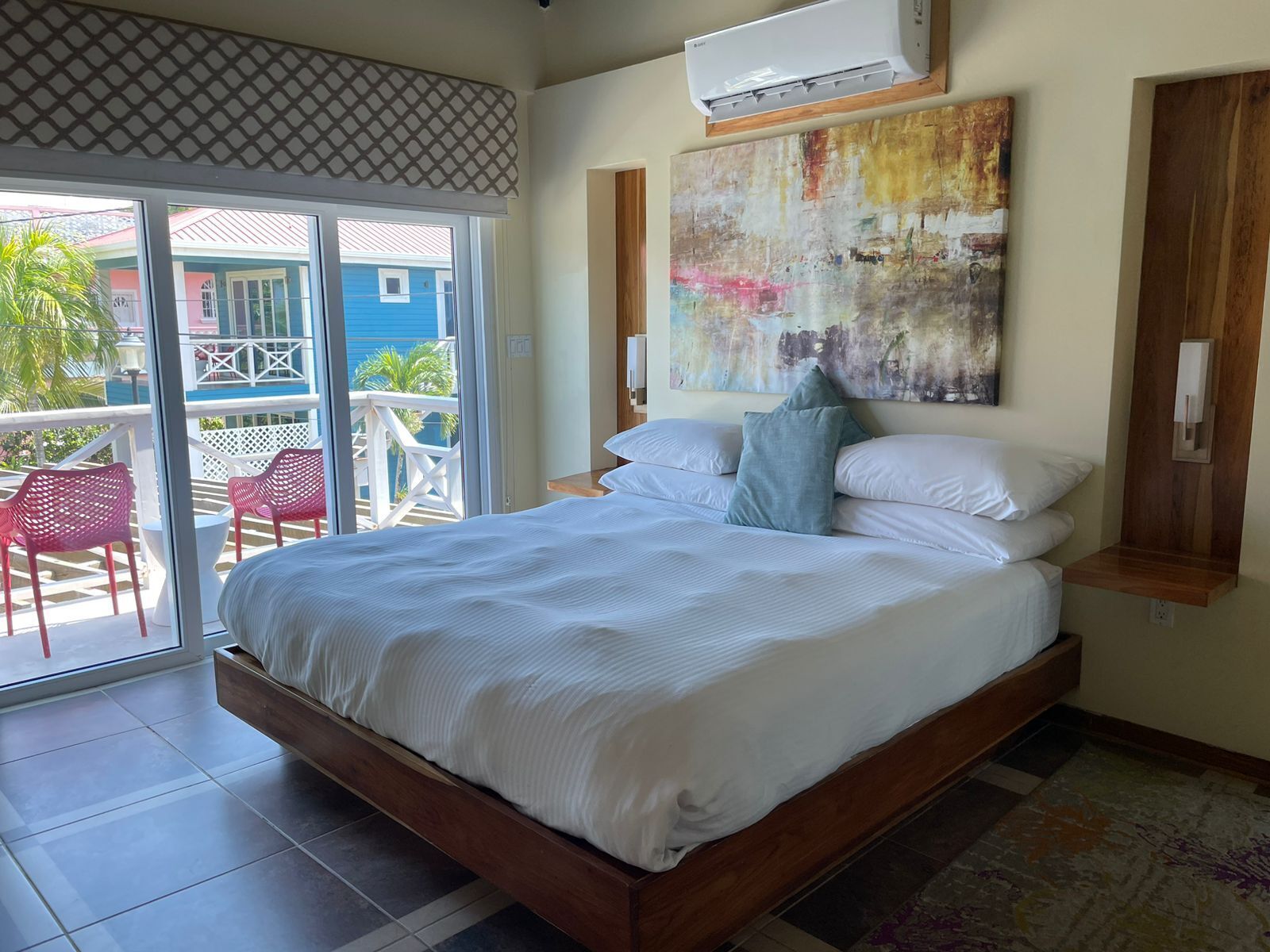 A bedroom with a large bed, a sliding glass door leading to a balcony, and a colorful abstract painting on the wall.