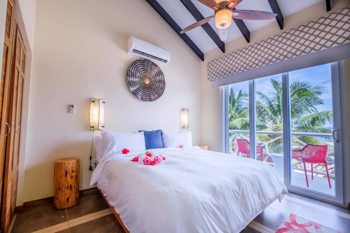 A hotel room with a large bed, two wall-mounted lamps, an air conditioner, and a view of palm trees through large windows.