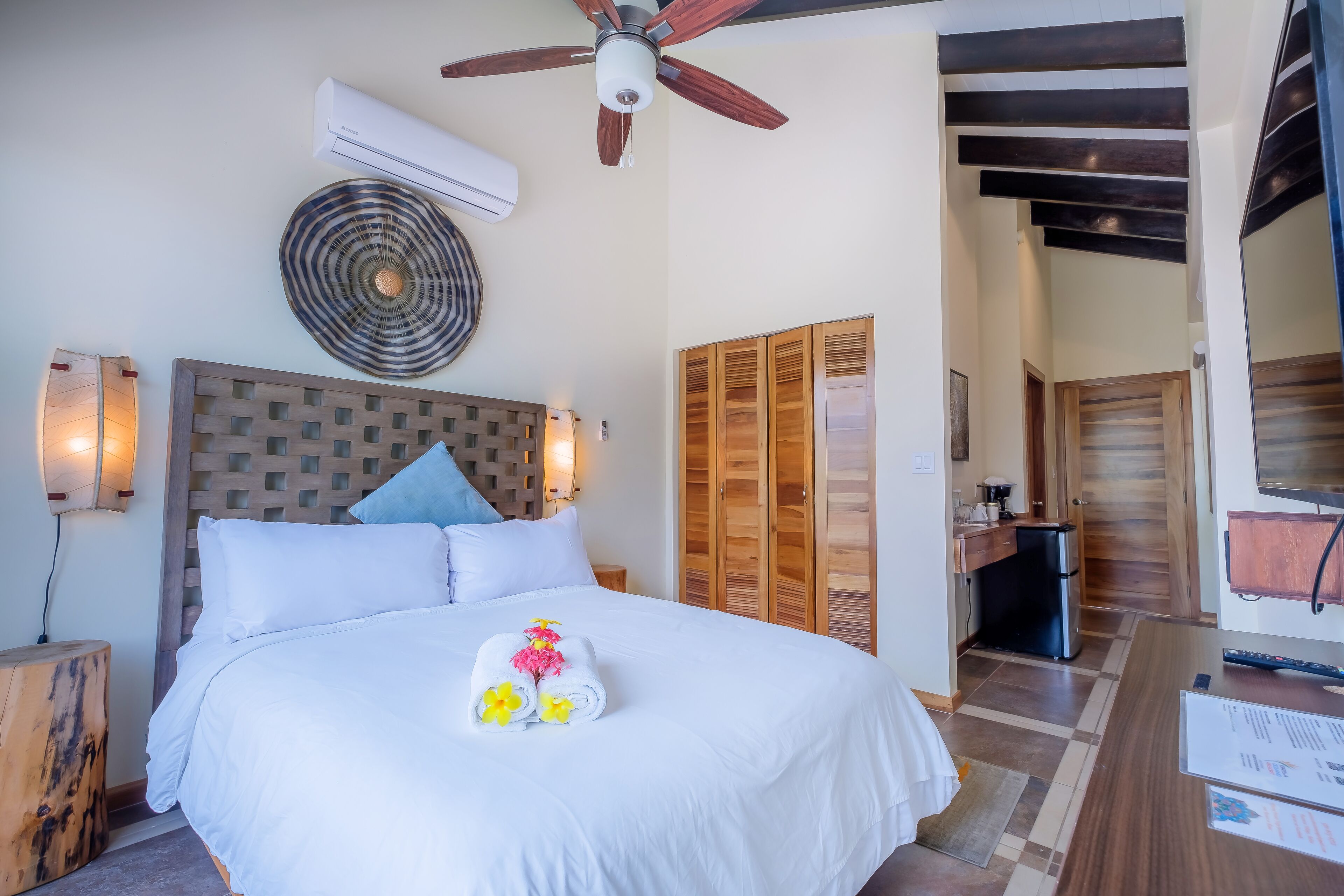 Een hotelkamer met een bed, een plafondventilator, een airconditioningapparaat, een houten garderobe, een kleine kitchenette en een houten nachtkastje.