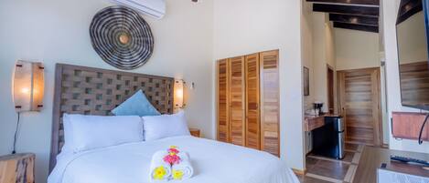 A hotel room with a bed, a ceiling fan, an air conditioning unit, a wooden wardrobe, a small kitchenette, and a wooden side table.