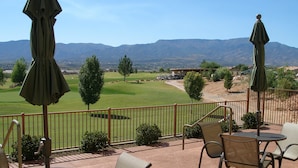 Um pátio externo com mesas e cadeiras redondas, vista para um campo de golfe e montanhas ao fundo.