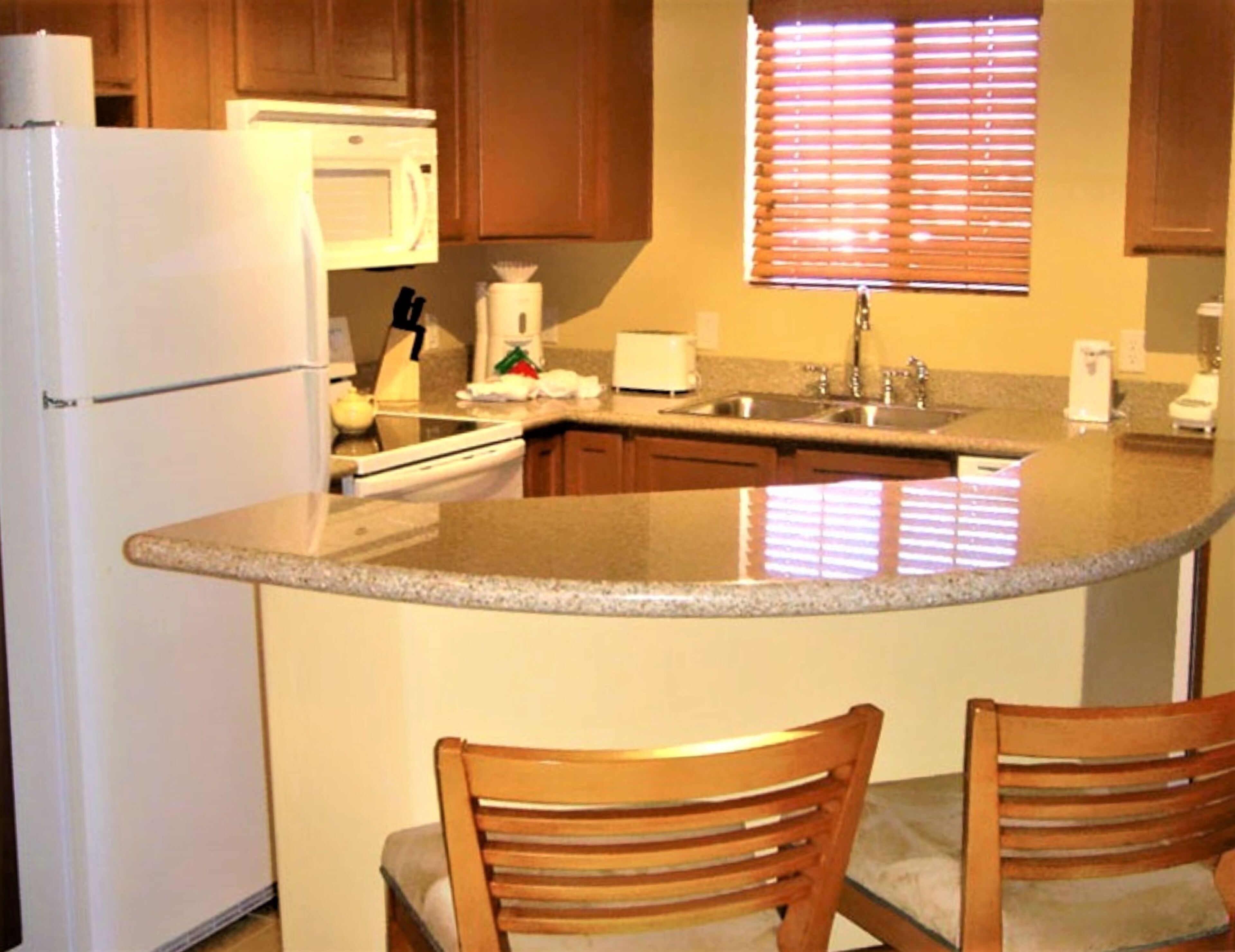 Una cocina con encimera de granito, armarios de madera, refrigerador, microondas, pileta y dos sillas de madera.