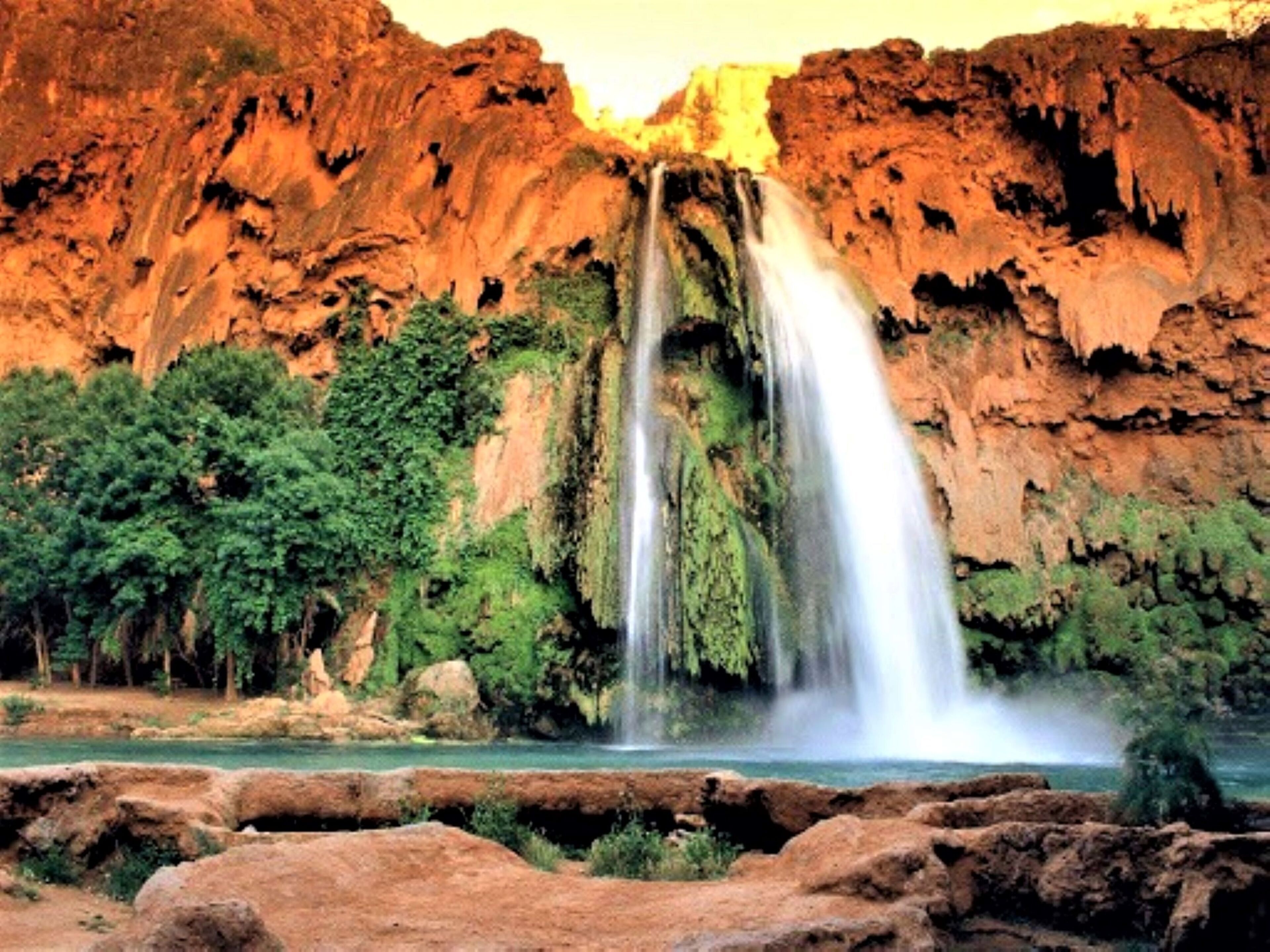 Uma cascata desce por um penhasco rochoso em direção a um rio, cercado por vegetação.