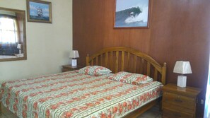 A bedroom with a wooden headboard, floral bedspread, two bedside tables with lamps, and two framed pictures on the wall. - Punta Roca Surf Resort (La Libertad)
