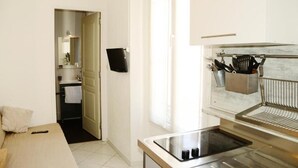 A modern kitchen with a built-in stove, a hanging utensil rack, and a view into the adjacent bathroom. - Hôtel L'Oustaou (Le Lavandou)