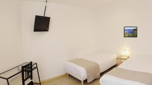 A small, simple room with two beds, a desk, and a TV mounted on the wall. - Hôtel L'Oustaou (Le Lavandou)