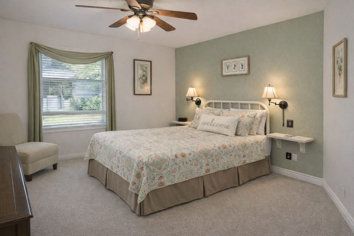 A bedroom with a bed, a ceiling fan, a window with curtains, wall-mounted lamps, and framed pictures.