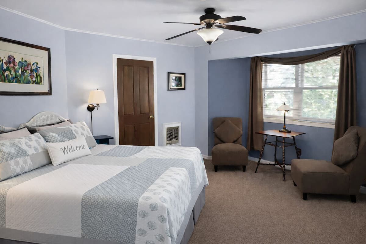 A bedroom with a bed, a chair, a table, a ceiling fan, a door, and a window.