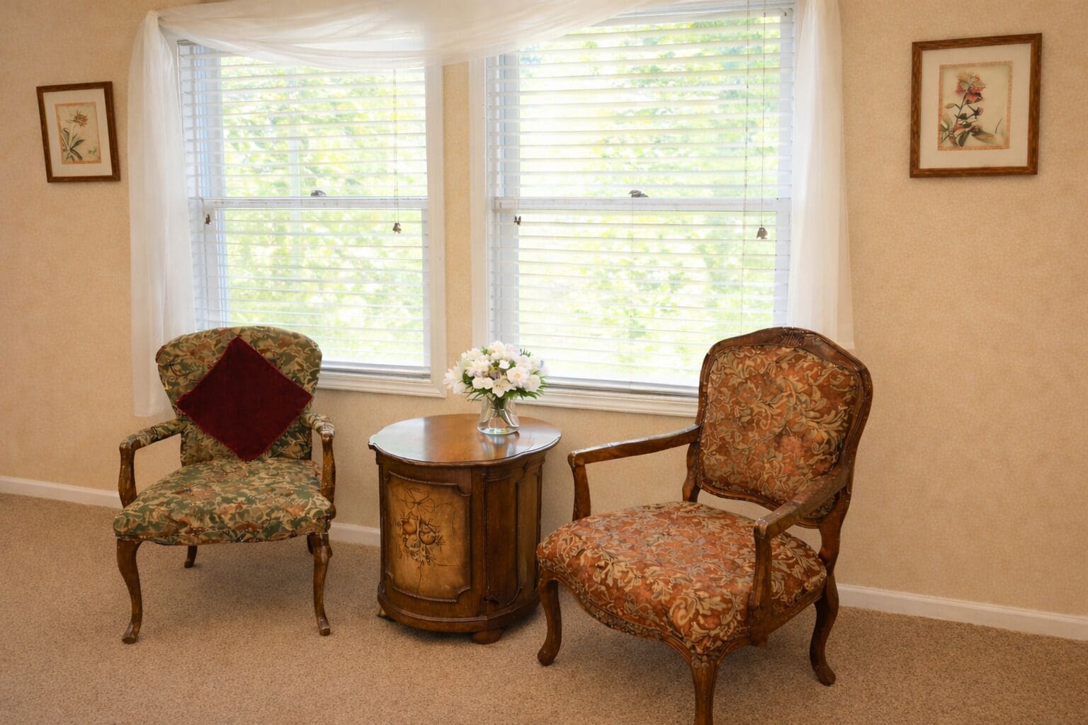 Dos sillones tapizados con estampados florales, una mesita redonda de madera con un jarrón de flores y una ventana con persianas.