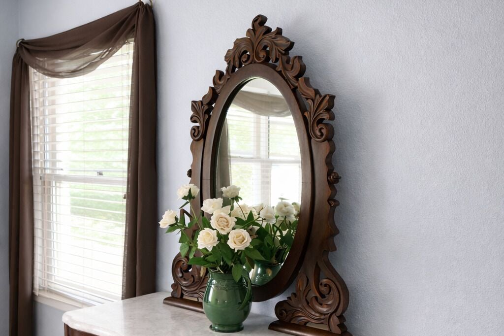A mirror with an ornate frame, a window with blinds, and a vase with white flowers on a table.