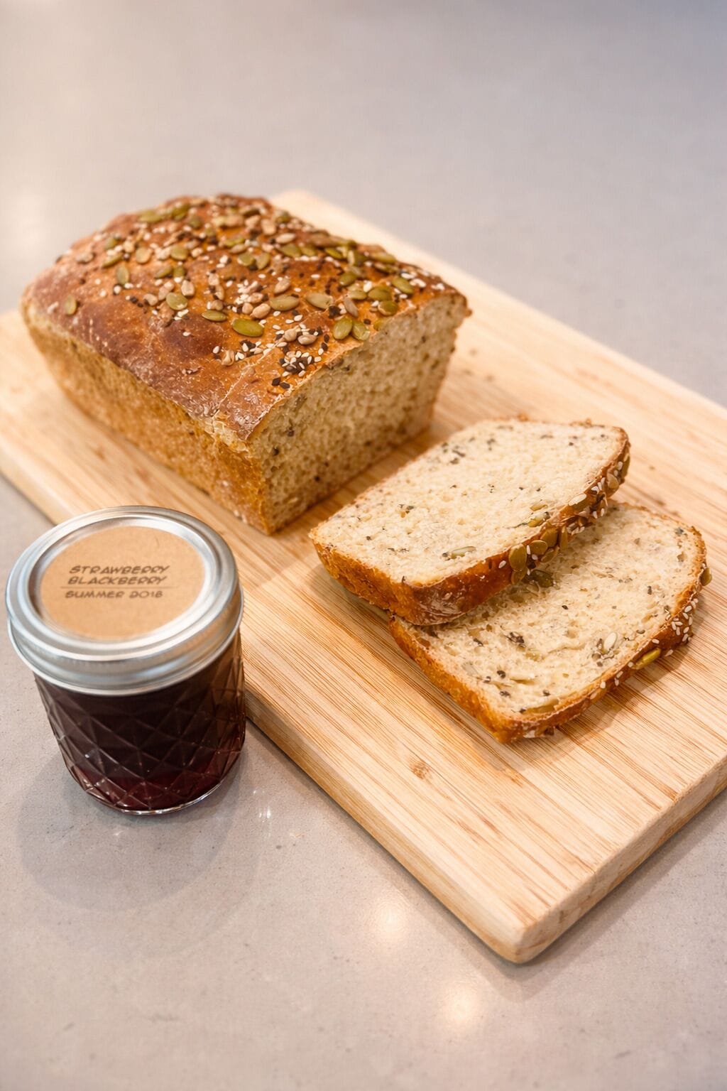 Una hogaza de pan con semillas, rebanadas de pan y un frasco de mermelada sobre una tabla de madera.