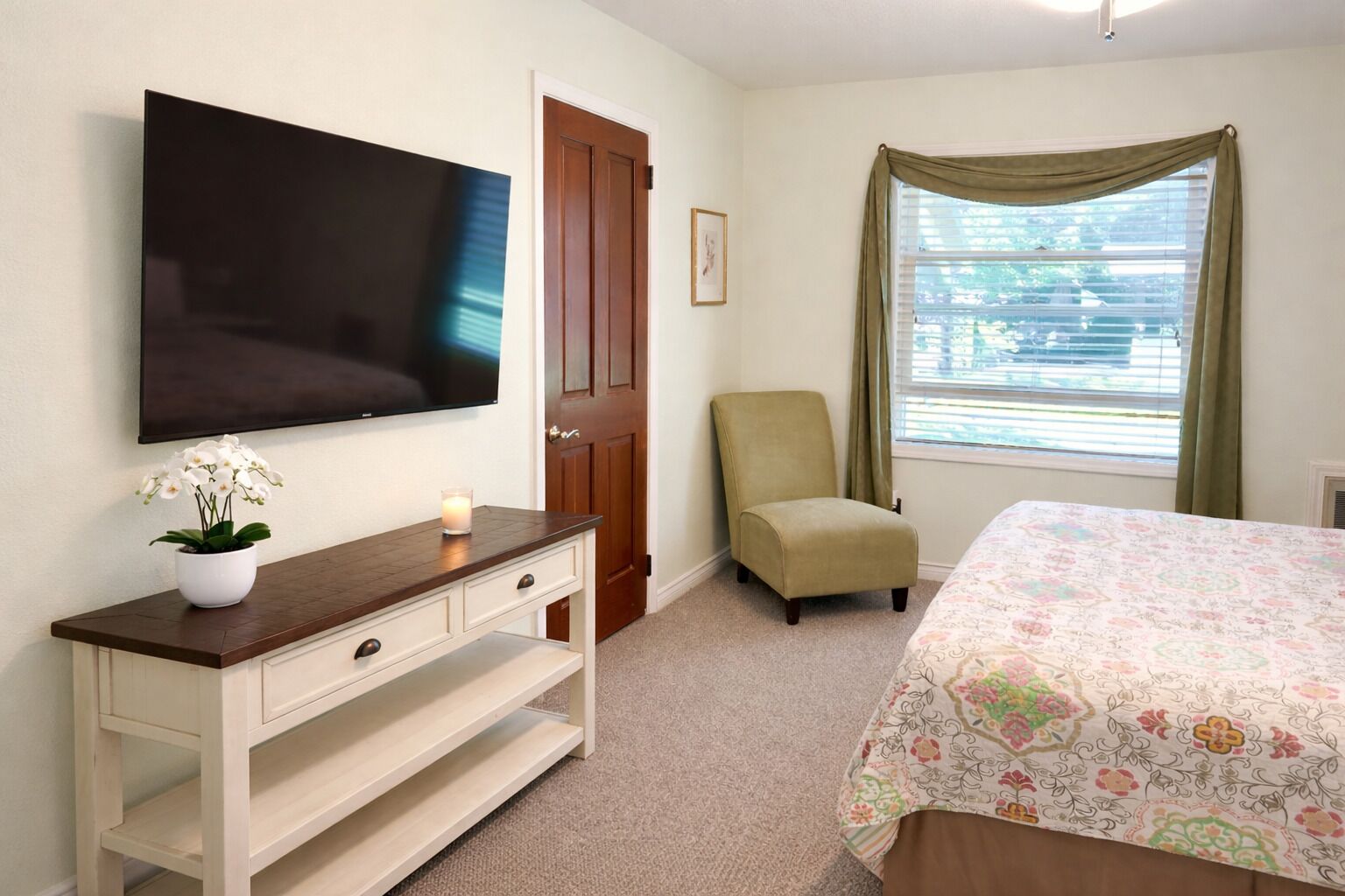 Quarto com cama, televisão na parede, cadeira, escrivaninha com planta e janela com cortinas.