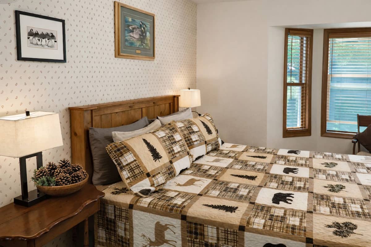 A bedroom with a wooden headboard, a quilted bedspread, two bedside lamps, a framed picture on the wall, and a window with blinds.