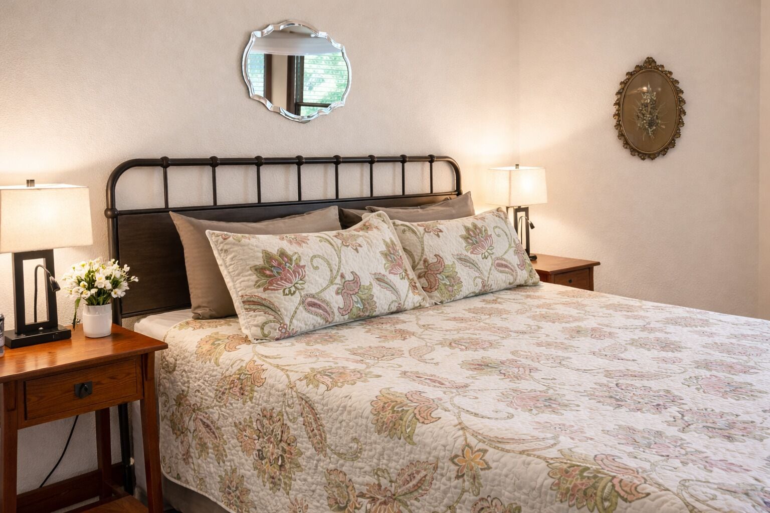 A neatly made bed with floral bedding, two bedside lamps, a bedside table with a vase of flowers, and a mirror on the wall.
