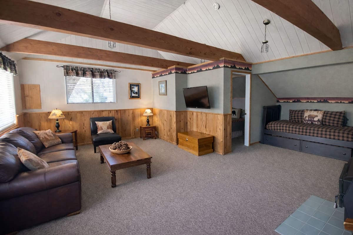 A cozy living room with a leather sofa, a wooden coffee table, a TV mounted on the wall, and a bed with checkered bedding.