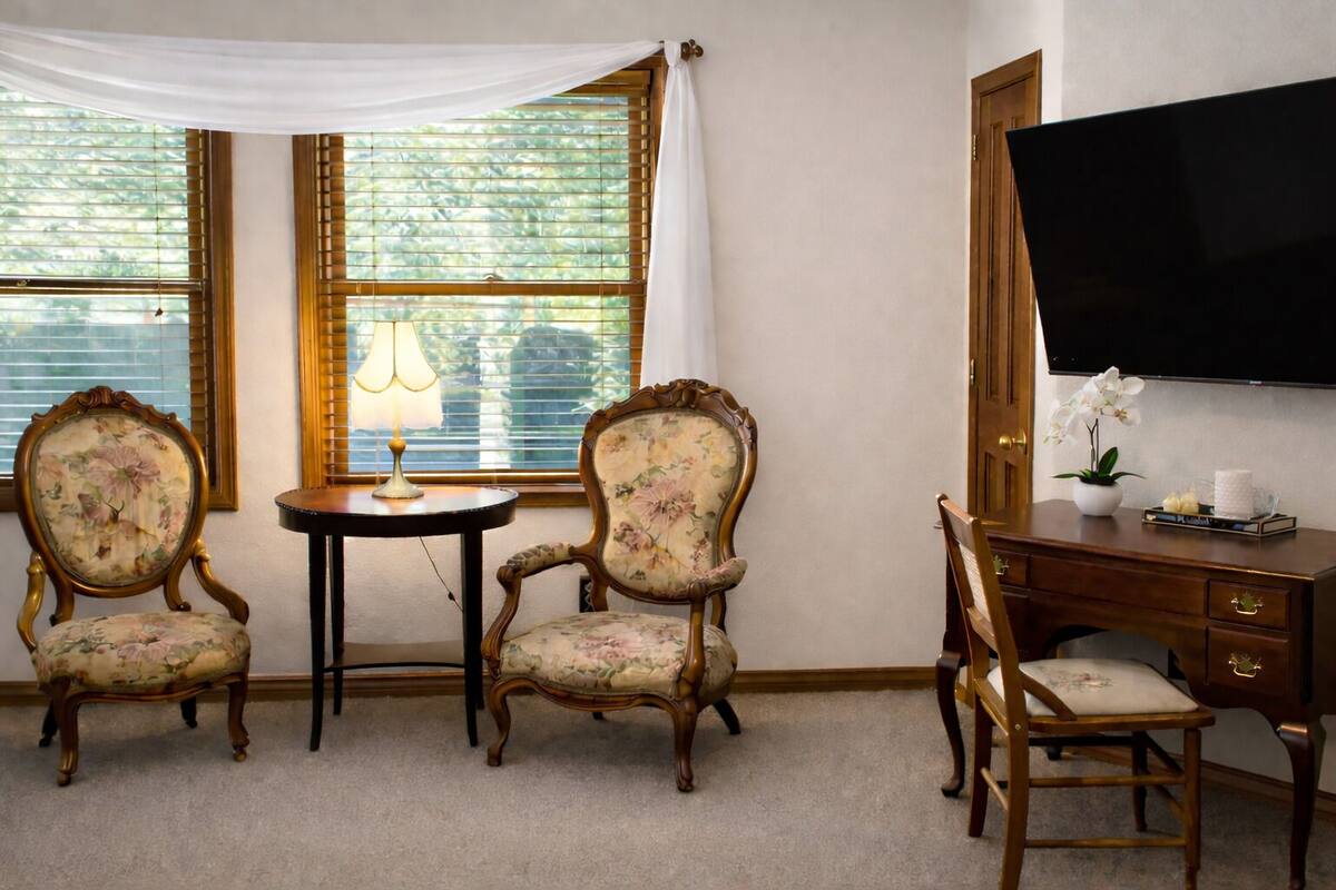 A room with two upholstered chairs, a small round table with a lamp, a wooden desk with a flat-screen TV, and a large window with blinds.