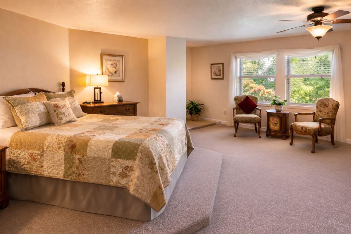 A bedroom with a large bed, a nightstand, a lamp, two chairs, a window with a view, and a ceiling fan.