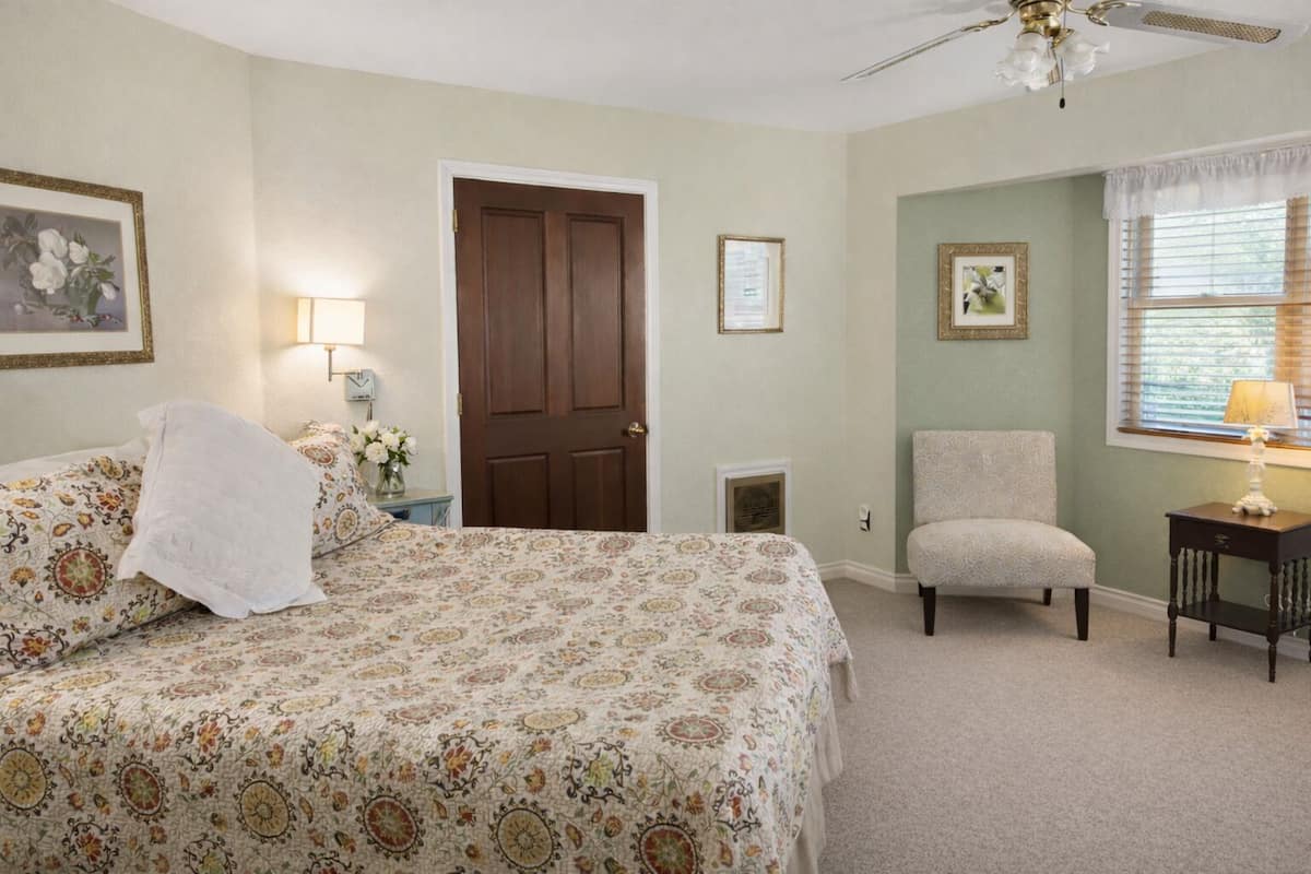 A bedroom with a bed, a chair, a desk, a door, and a window with blinds.