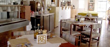 A cozy dining area with wooden tables and chairs, a kitchen in the background, and a large window providing natural light.