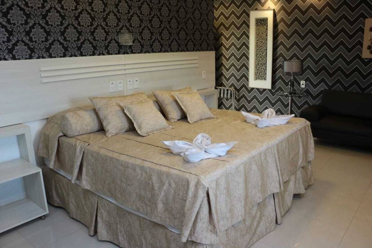 Una cama bien tendida con cubrecama y almohadas color beige, una mesita de noche, y una pared con un espejo decorativo y papel tapiz estampado.
