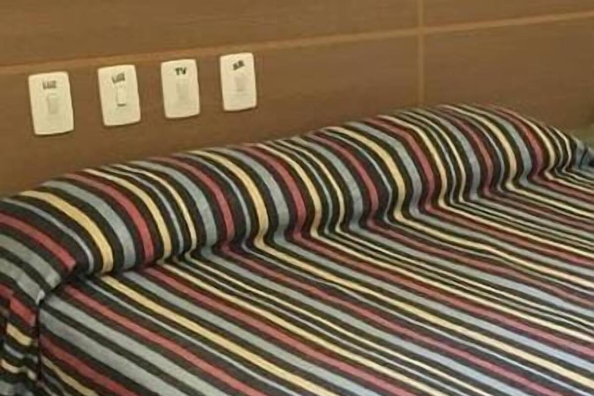 Una cama con una colcha a rayas y una pared con cuatro interruptores etiquetados como “Luz”, “TV”, “TV” y “AP”.