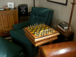A chessboard on a wooden table in a room with a green armchair, a wooden cabinet, and a radio.
