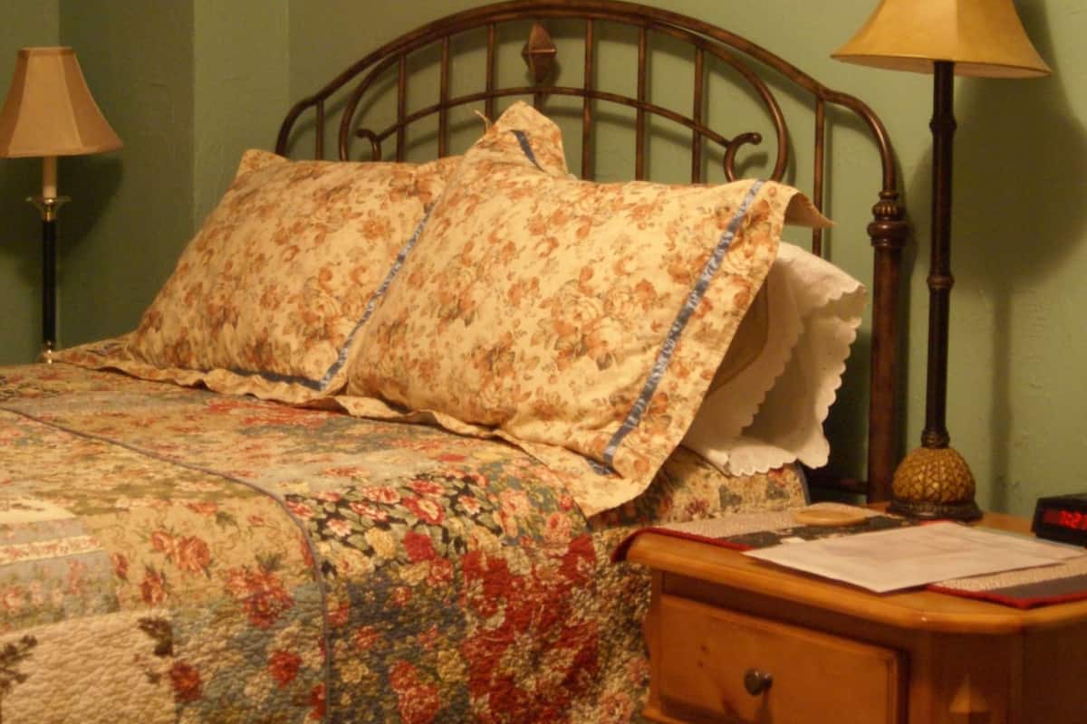 A bedroom with a floral bedspread, a wooden nightstand with a lamp, and a painting on the wall.