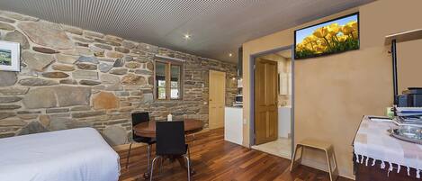 A room with a stone wall, a wooden dining table with chairs, a bed, and a television on the wall.