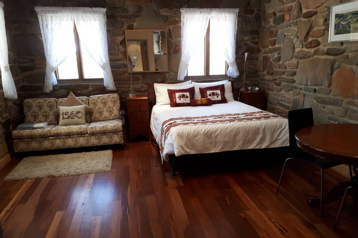 A cozy room with a wooden floor, a bed with white bedding and decorative pillows, a sofa with patterned upholstery, a small table, and a stone wall.