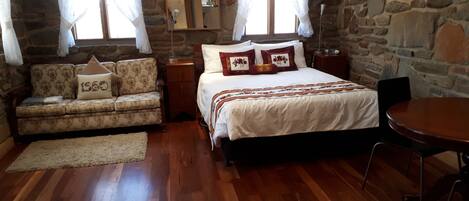 A cozy room with a wooden floor, a bed with white bedding and decorative pillows, a sofa with patterned upholstery, a small table, and a stone wall.