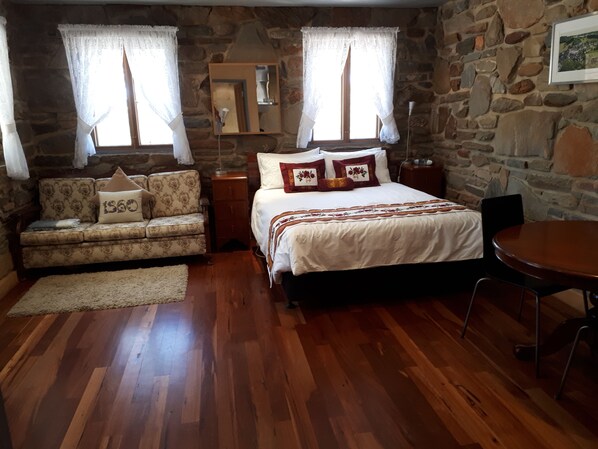 A cozy room with a wooden floor, a bed with white bedding and decorative pillows, a sofa with patterned upholstery, a small table, and a stone wall. - 1860 Wine Country Cottages (Springton)