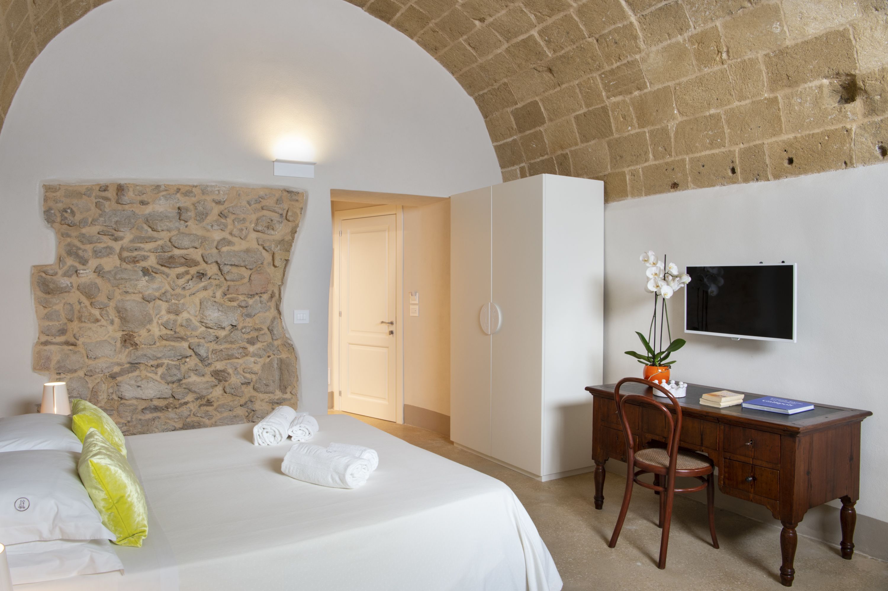 A bedroom with a stone wall, a bed with white linens, a desk with a chair, and a television on the wall.
