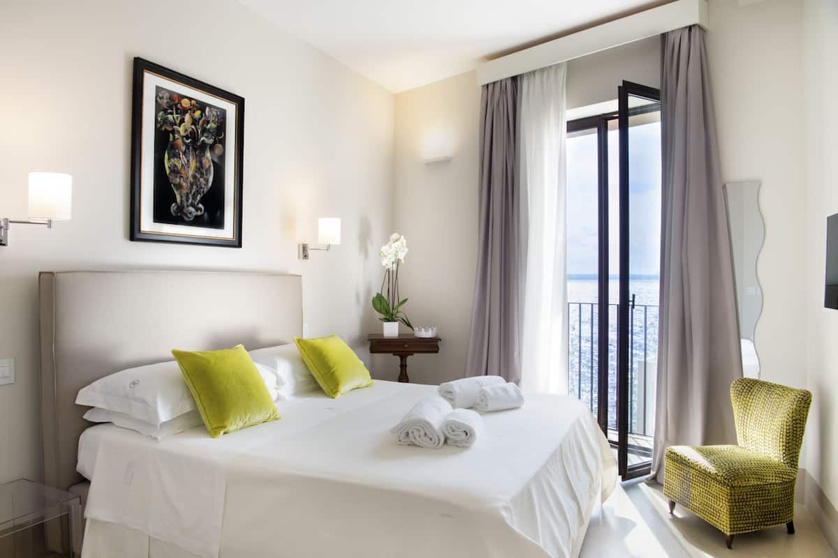 Habitación de hotel con una cama grande, dos almohadas, una mesita con una planta, una silla y vistas a la ciudad.