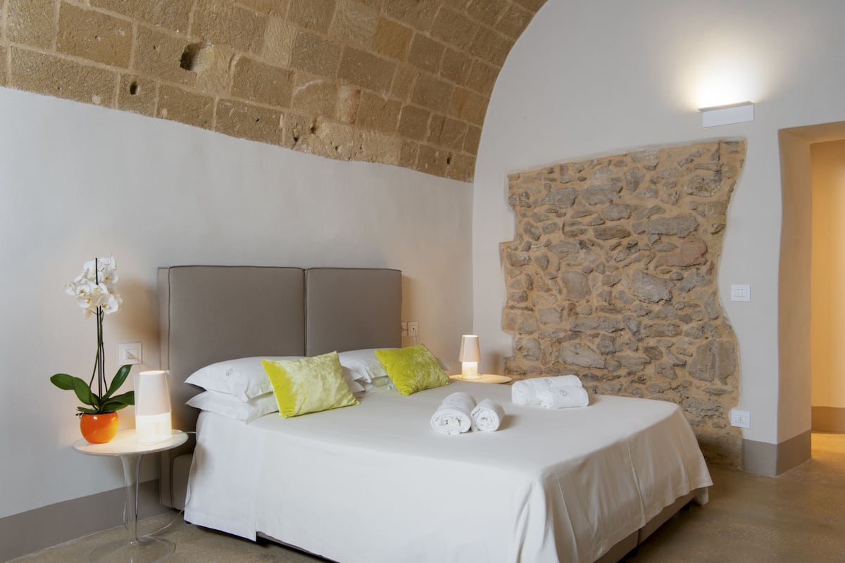 Un dormitorio con pared de piedra, cama con ropa blanca, mesita de noche con lámpara y una planta en maceta.