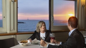 Une table à manger avec vue sur le coucher de soleil sur l’océan, une lampe et une personne tenant un verre de vin.
