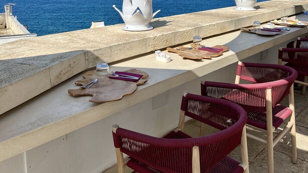 Une terrasse en bord de mer offrant une vue sur l’océan, avec une table, des chaises et des vases blancs décoratifs.