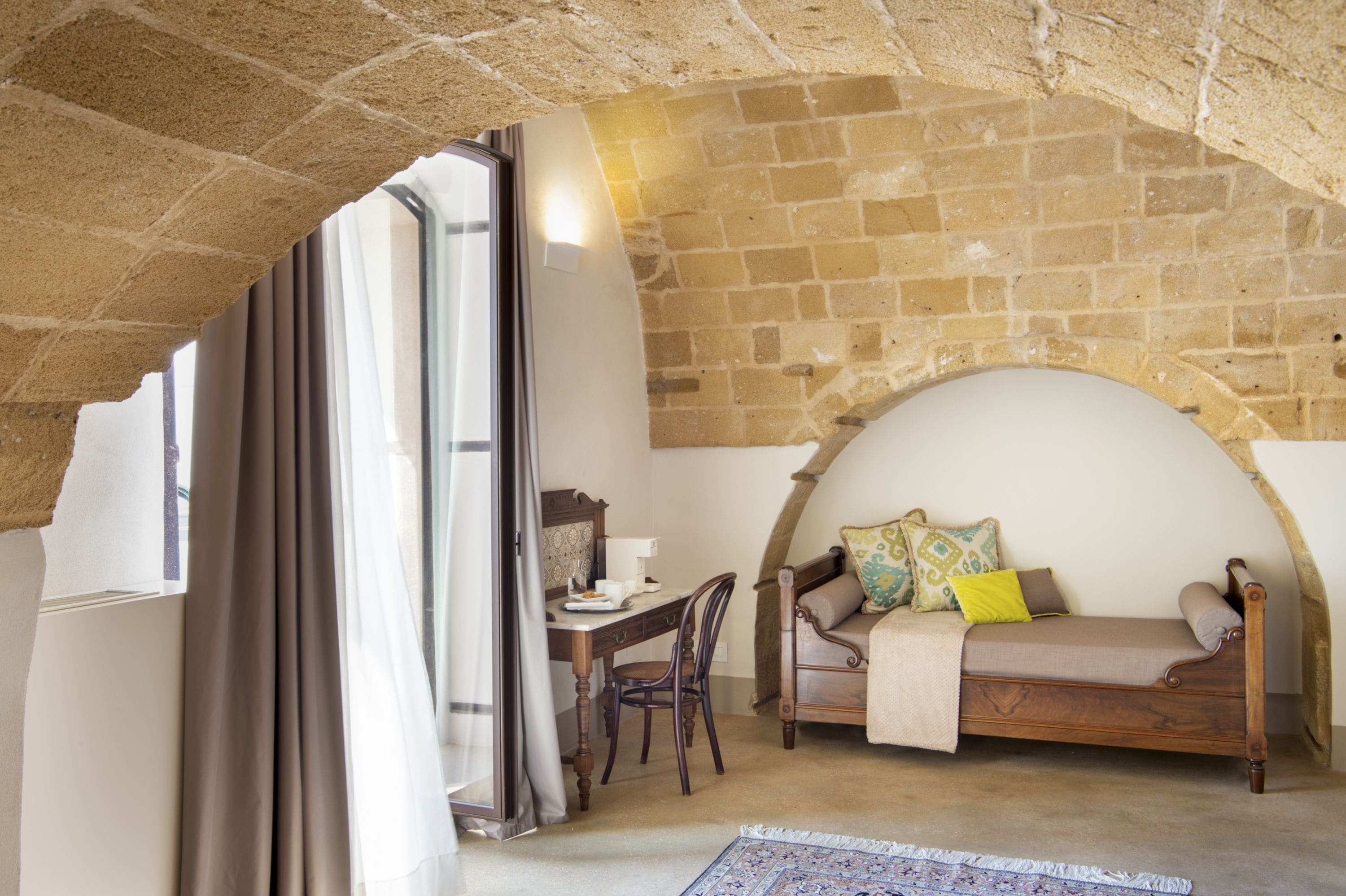 A cozy room with a stone wall, a wooden bed, a desk with a chair, and a window with curtains.