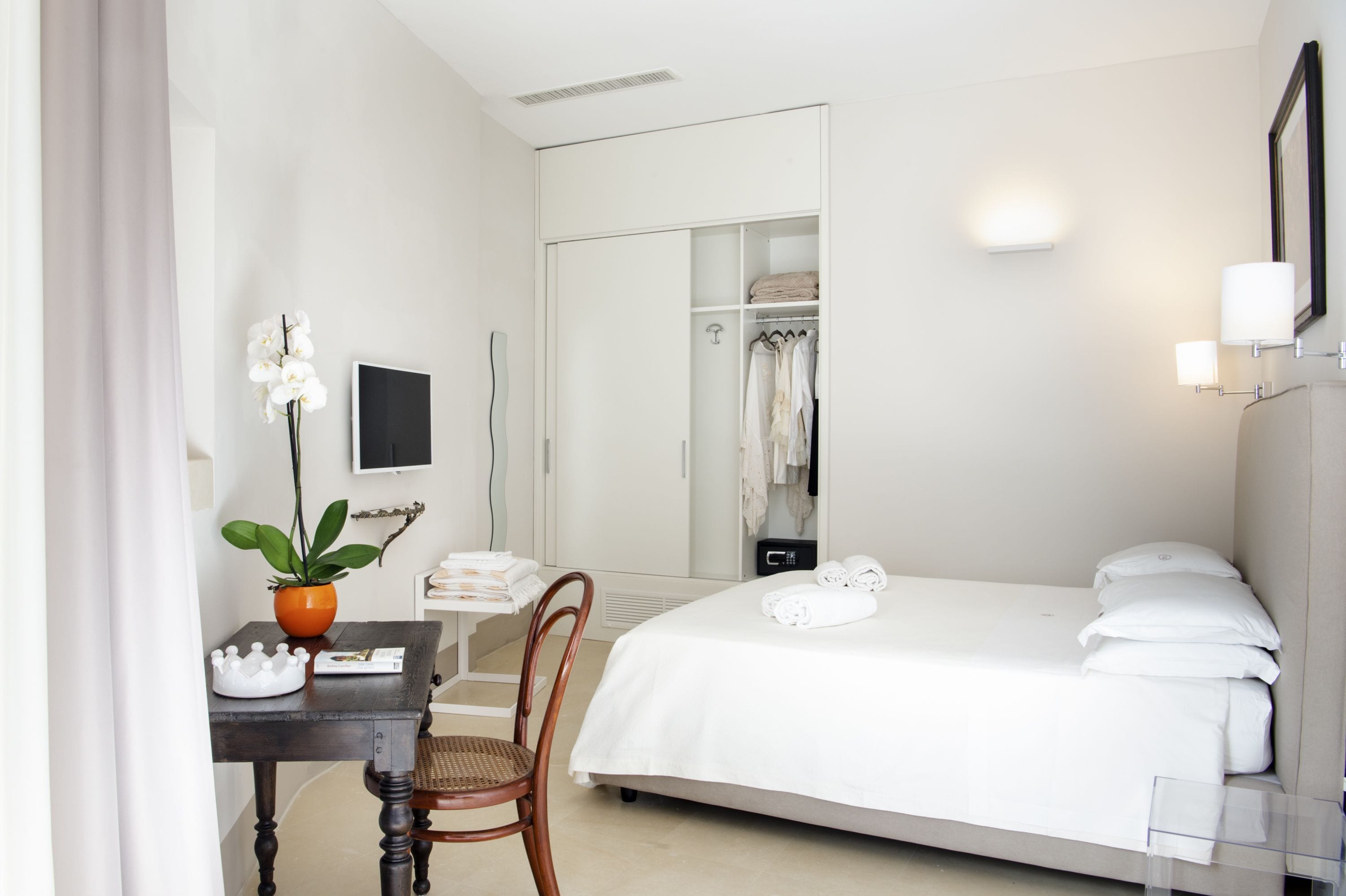 A hotel room with a bed, a desk with a chair, a potted plant, a TV, and a closet.