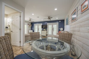 Un salon moderne avec une table basse en verre, des meubles en osier, un ventilateur de plafond et un téléviseur fixé au mur.